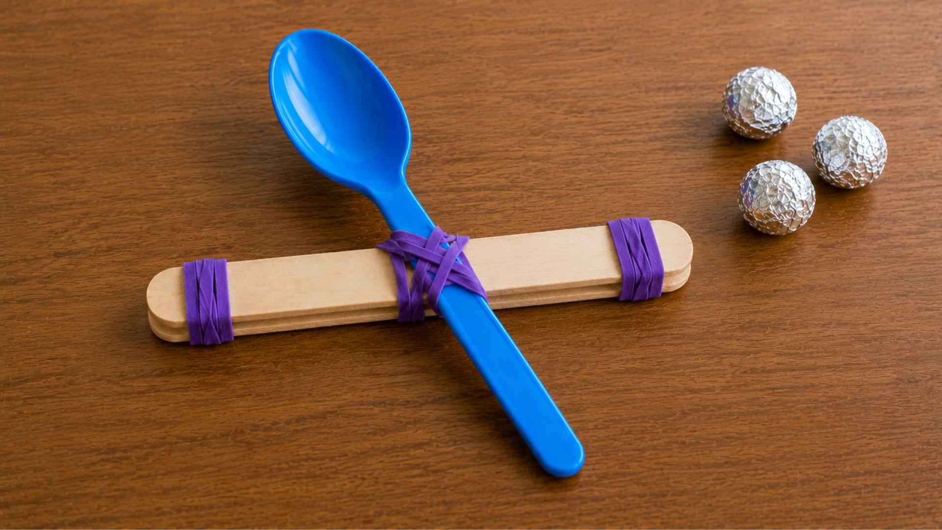 Completed popsicle stick catapult with blue spoon attached and foil balls nearby.