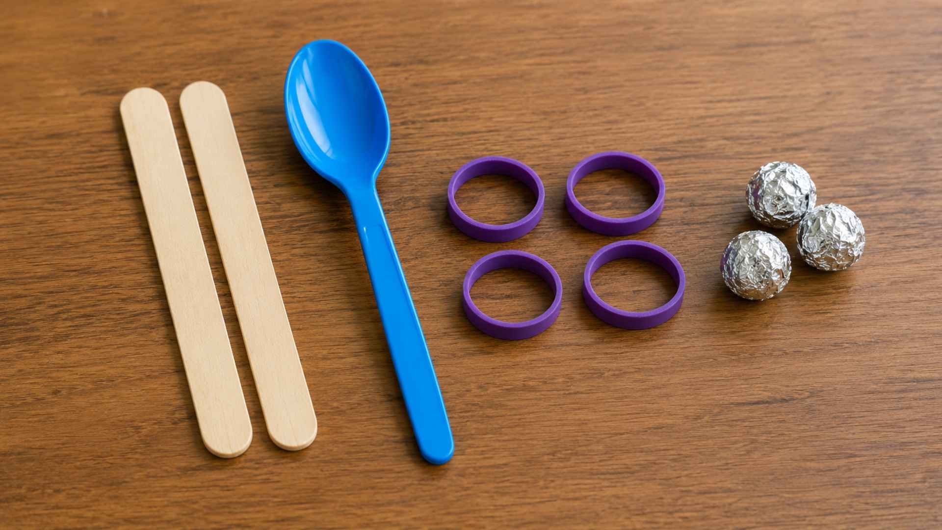 Popsicle sticks, blue spoon, purple rubber bands, and foil balls arranged on wooden table.
