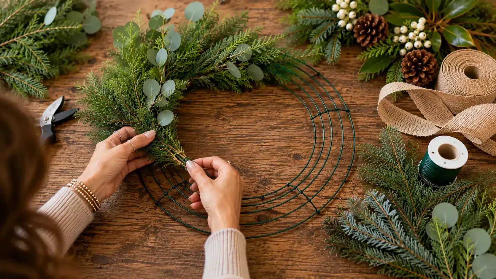 Person attaching fresh evergreen and eucalyptus stems to a wire wreath frame with floral wire