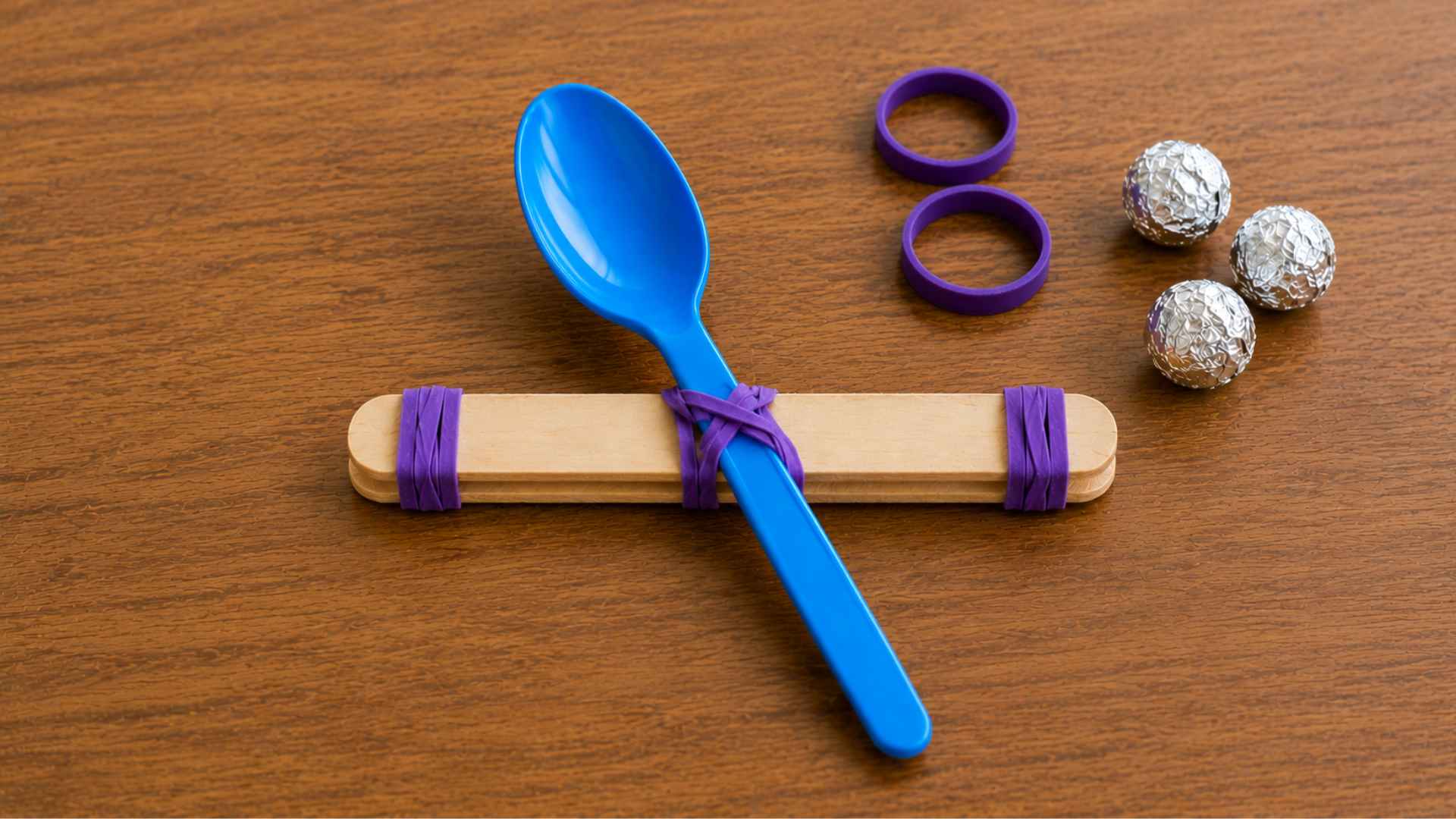 Purple rubber bands secure blue spoon to popsicle stick base on wooden tabletop.
