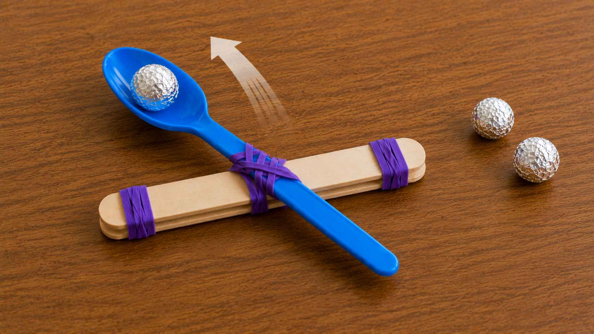 Loaded popsicle stick catapult ready to launch foil ball across tabletop.