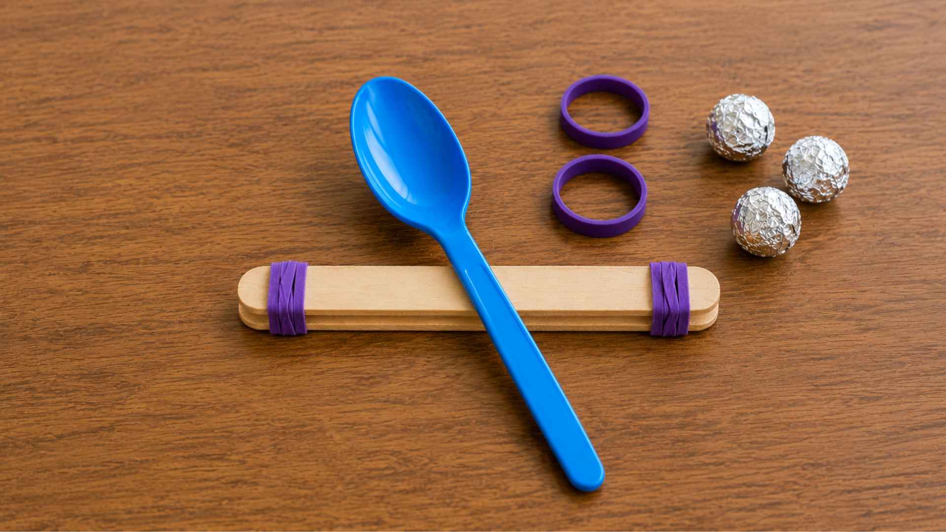 Blue spoon positioned diagonally across stacked popsicle sticks, forming catapult shape.