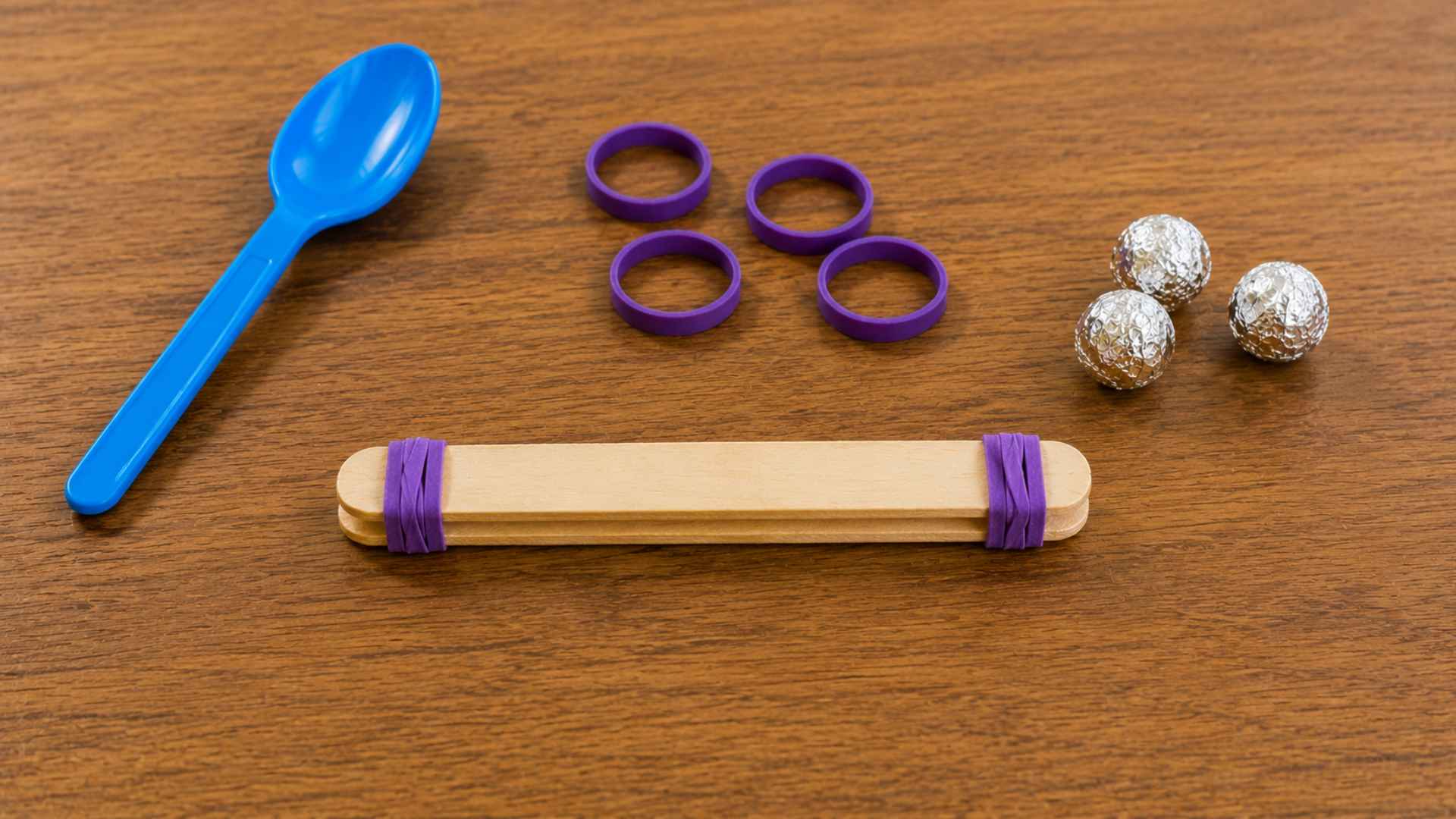 Two stacked popsicle sticks secured with purple rubber bands beside catapult supplies.