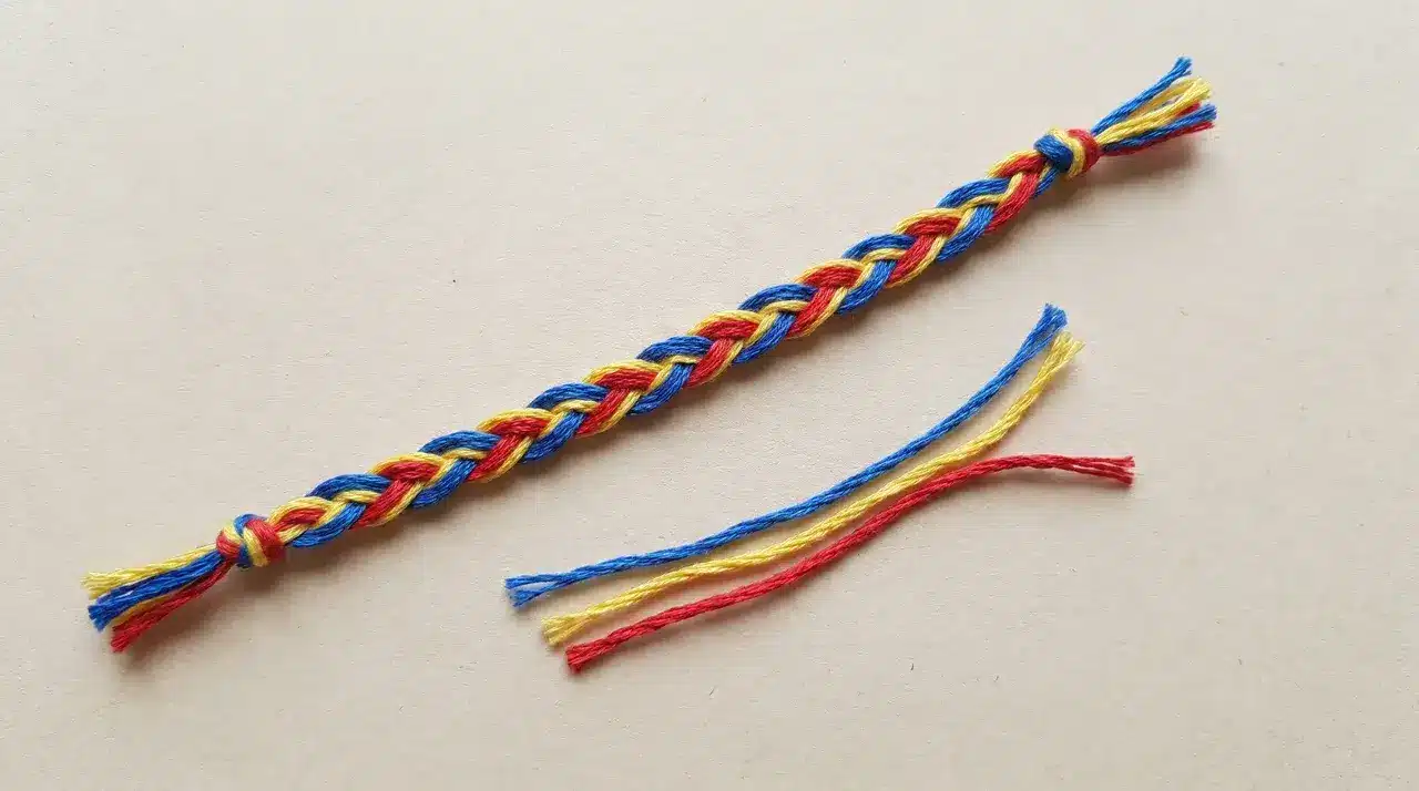 Braided three-color thread bracelet knotted at both ends on a light surface with loose thread strands beside it