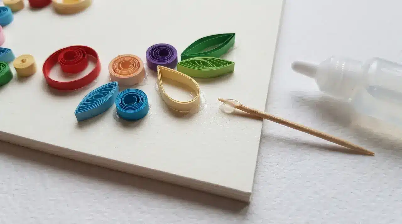 Quilled paper coils in circle, teardrop, and leaf shapes on white surface with toothpick and glue bottle