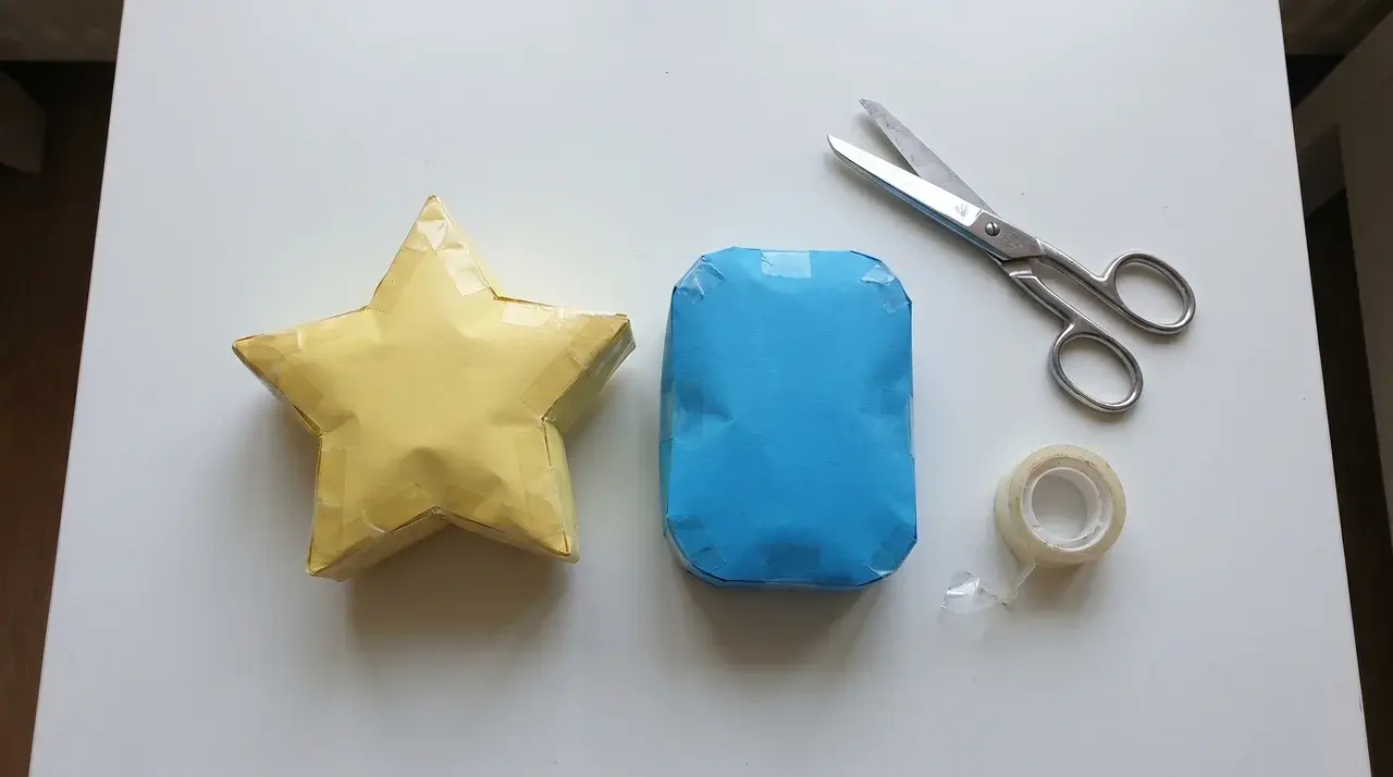 Two stuffed paper squishies in star and rectangle shapes on a white surface with scissors and tape nearby