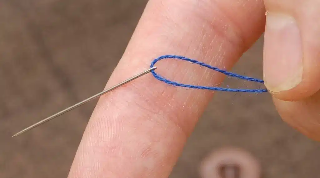 A close-up of fingers threading a blue loop of thread through a needle eye.
