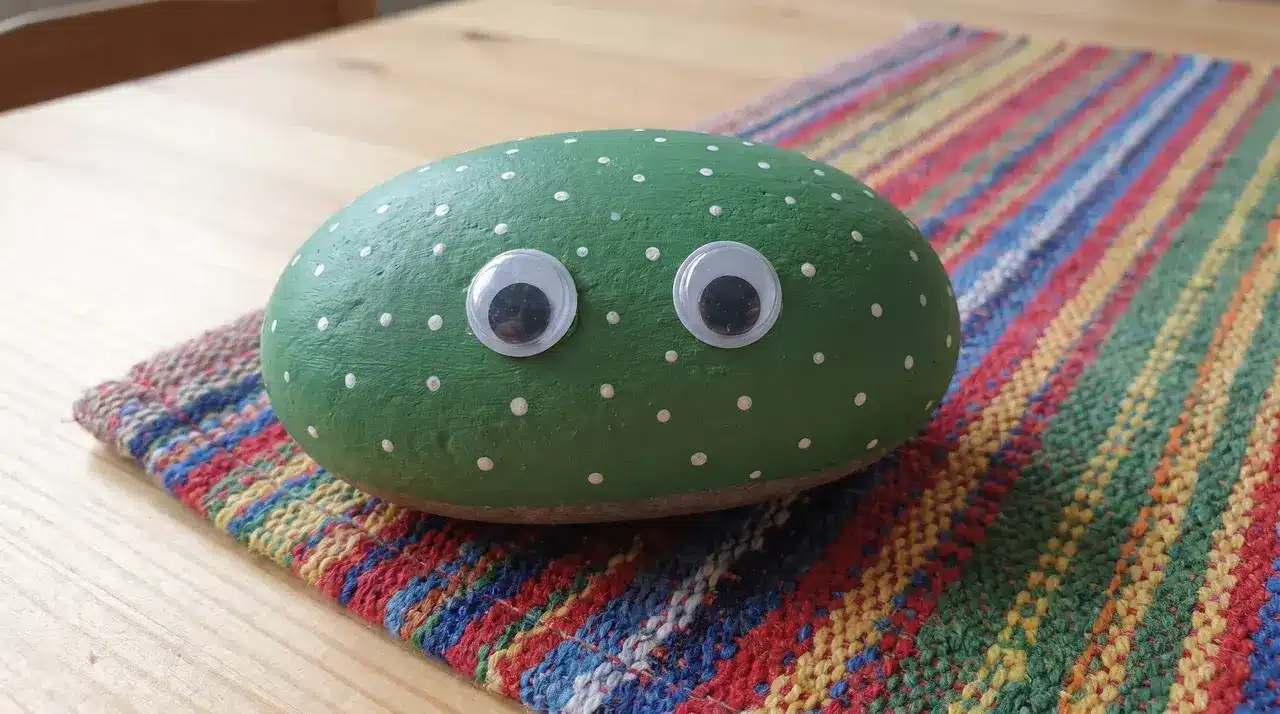 Green painted rock with white dots sitting on a colourful rug with googly eyes