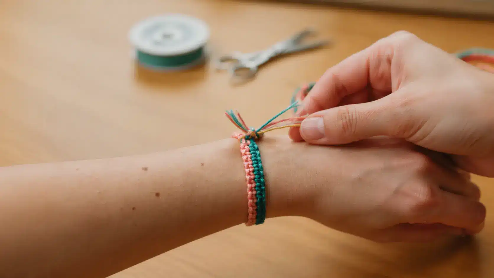 Hands adjusting the ends of a colorful braided friendship bracelet on a wrist over a wooden crafting surface.