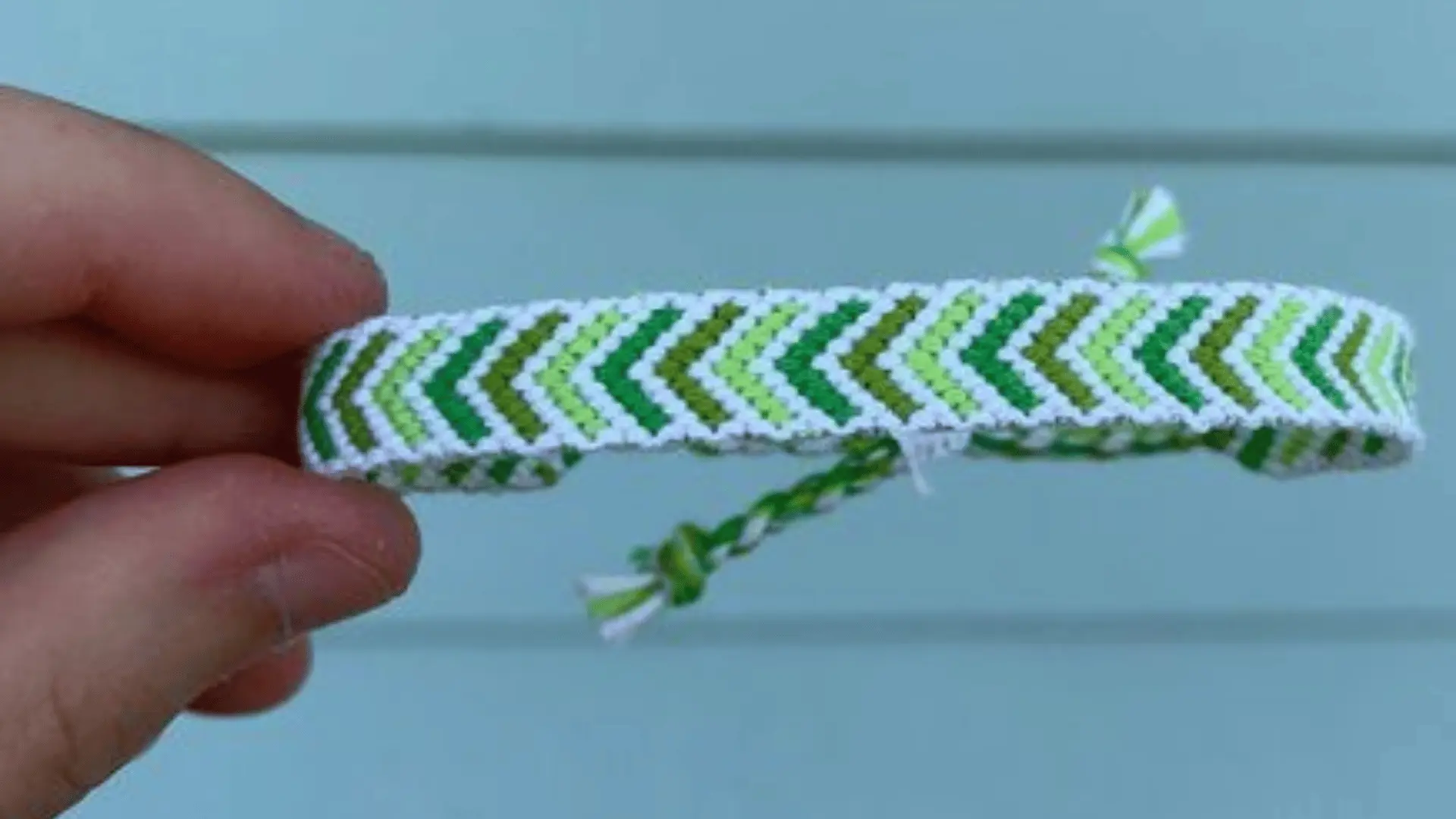 A hand holding a woven friendship bracelet with a green and white arrow or chevron pattern.