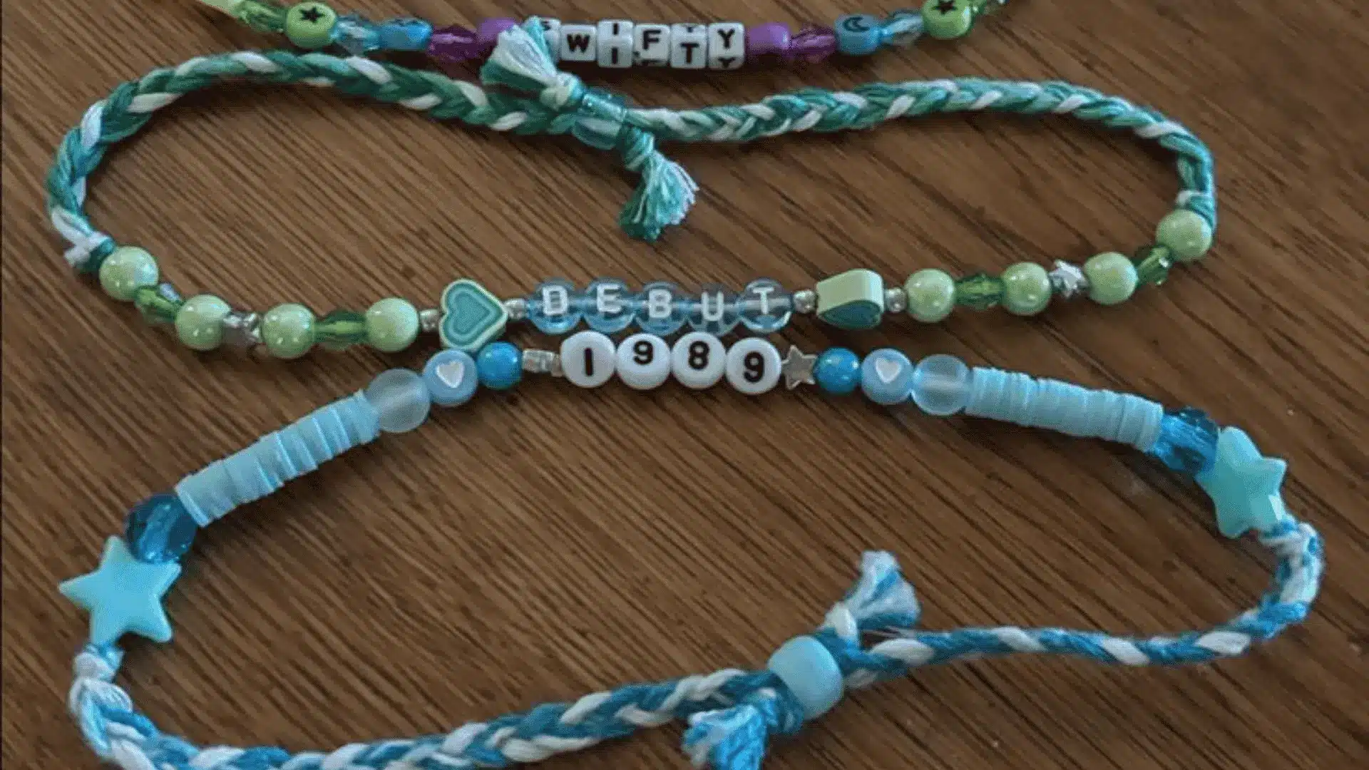Three handmade friendship bracelets featuring green and blue beads, braided string, and letter beads on a wooden surface.