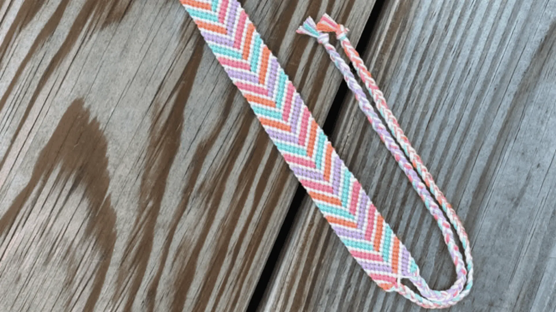 A woven friendship bracelet with a candy pastel chevron pattern in pink, orange, purple, and teal on a wooden deck.