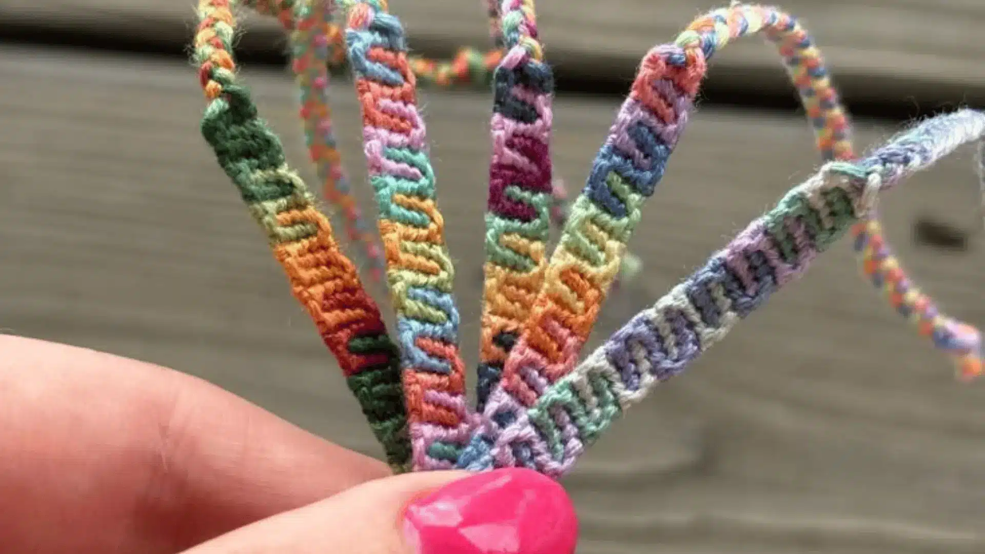 Five colorful candy wave bracelet strands held by a hand with pink nails against a blurry wooden deck.