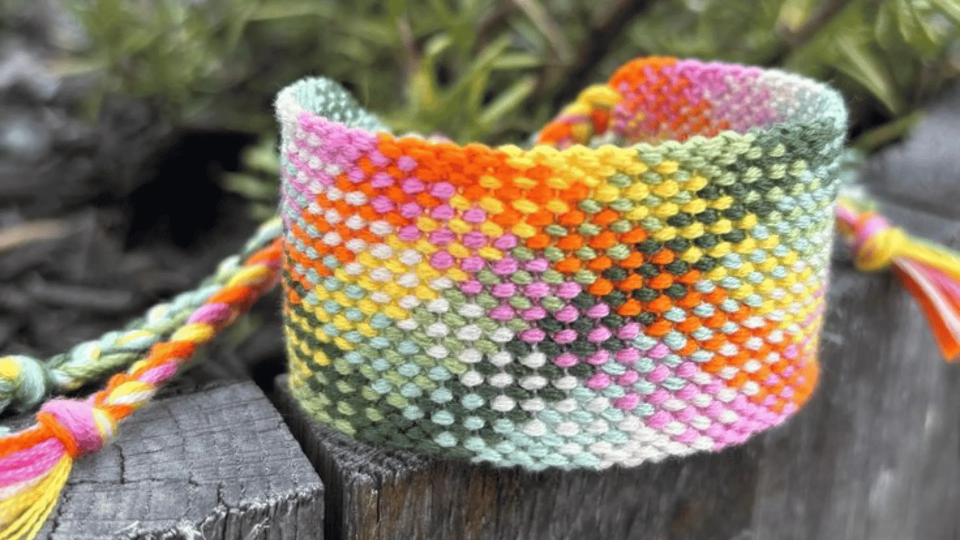 A chunky wide band woven friendship bracelet with a vibrant, multicolored checkered pattern resting on a wooden log.