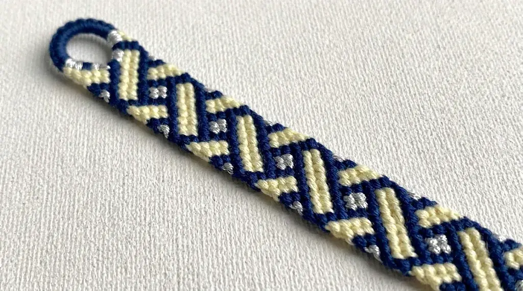 A woven friendship bracelet with a navy blue, cream, and silver criss-cross pattern resting on a white surface.