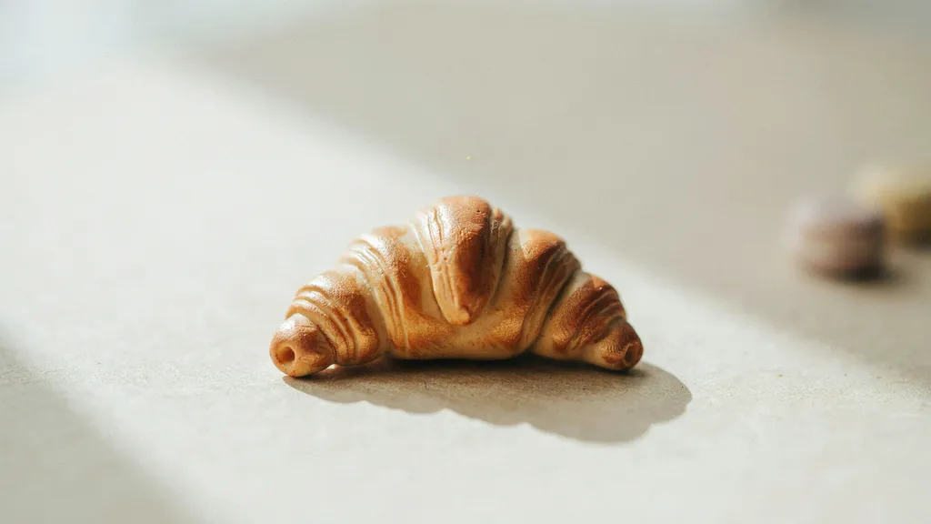 A handmade polymer clay croissant charm lying flat on a cream table surface with layered texture and warm golden baked tones under soft lighting.