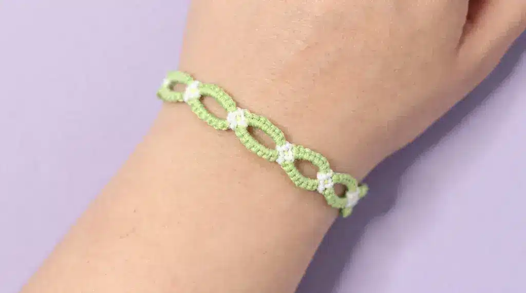 A woven Daisy Chain Bracelet with green loops and small white flowers worn on a wrist against a purple background.