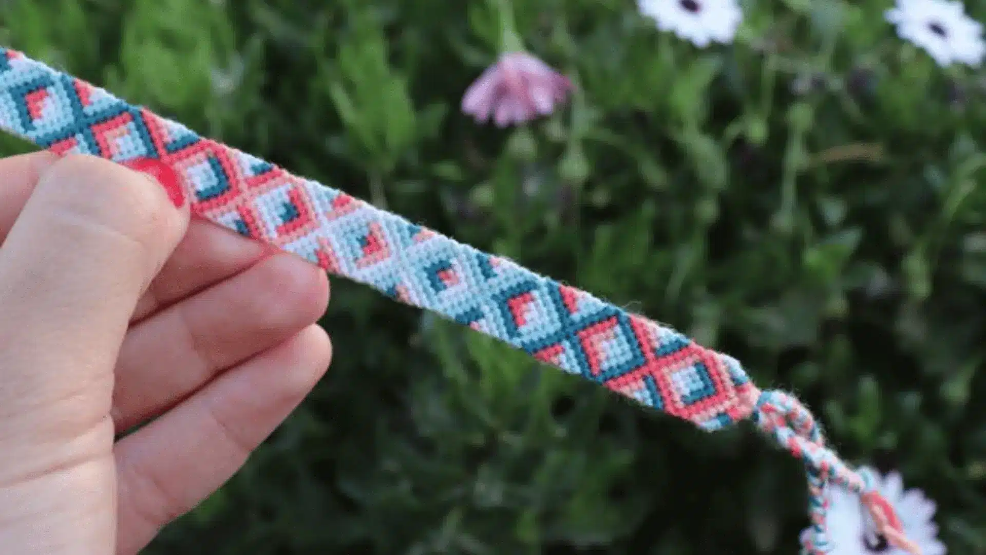 A hand holding a woven friendship bracelet with a colorful diamond chain pattern against a grassy background.