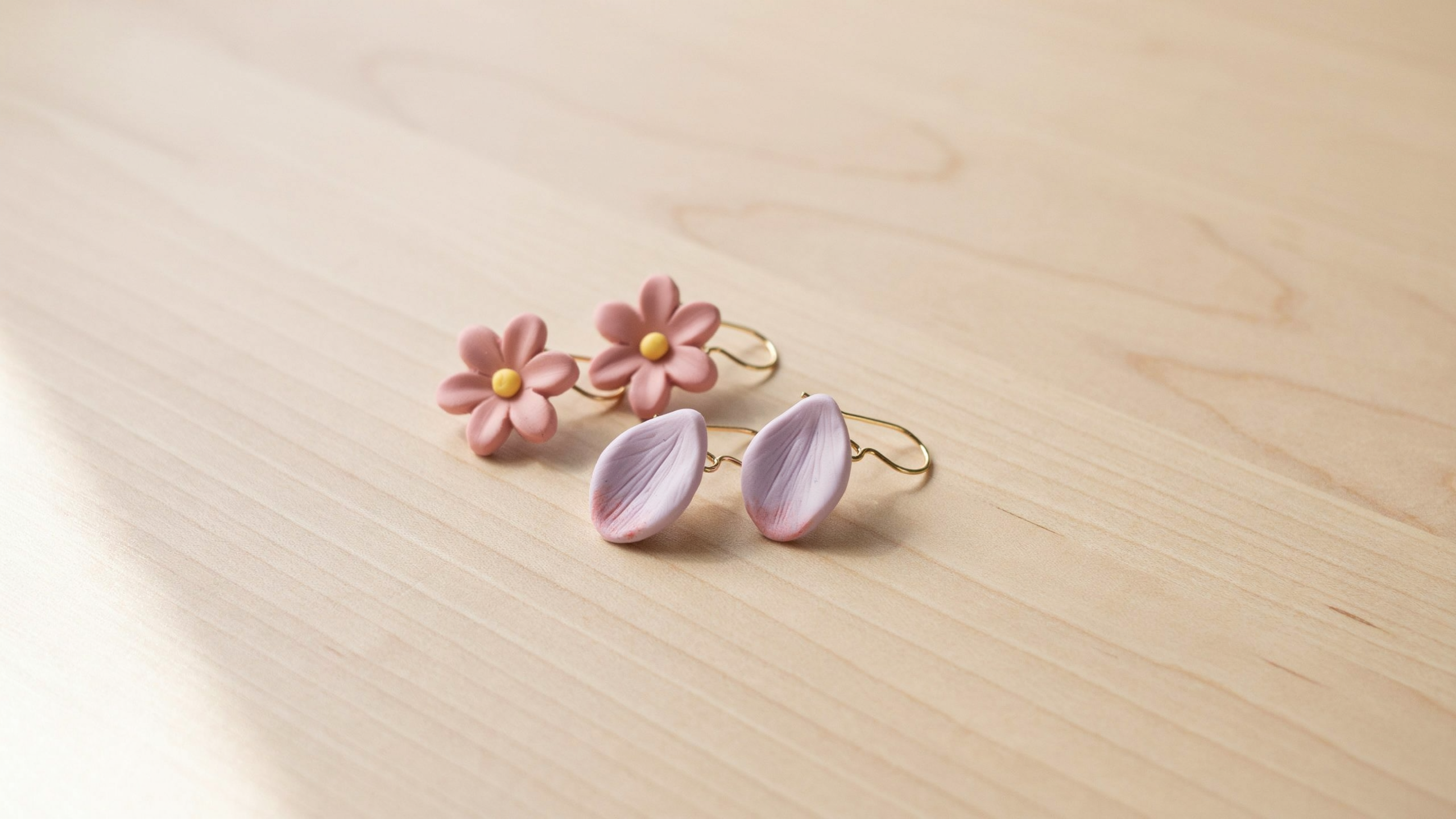 A pair of lightweight handmade floral clay earrings shaped like small flowers, attached to metal hooks, displayed neatly on a soft neutral surface.