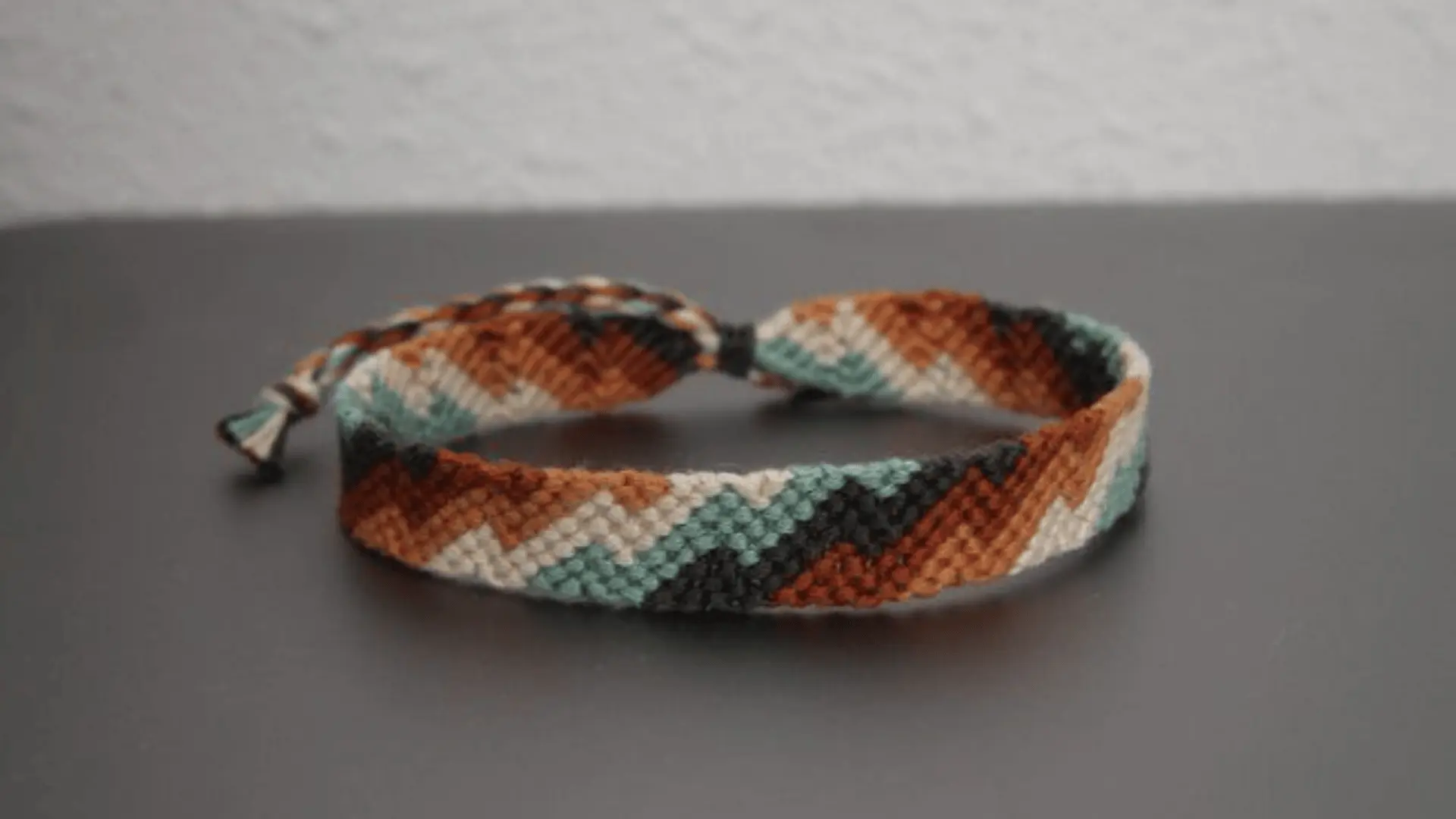 A woven friendship bracelet with a chevron pattern in earth tones like brown, cream, teal, and black.