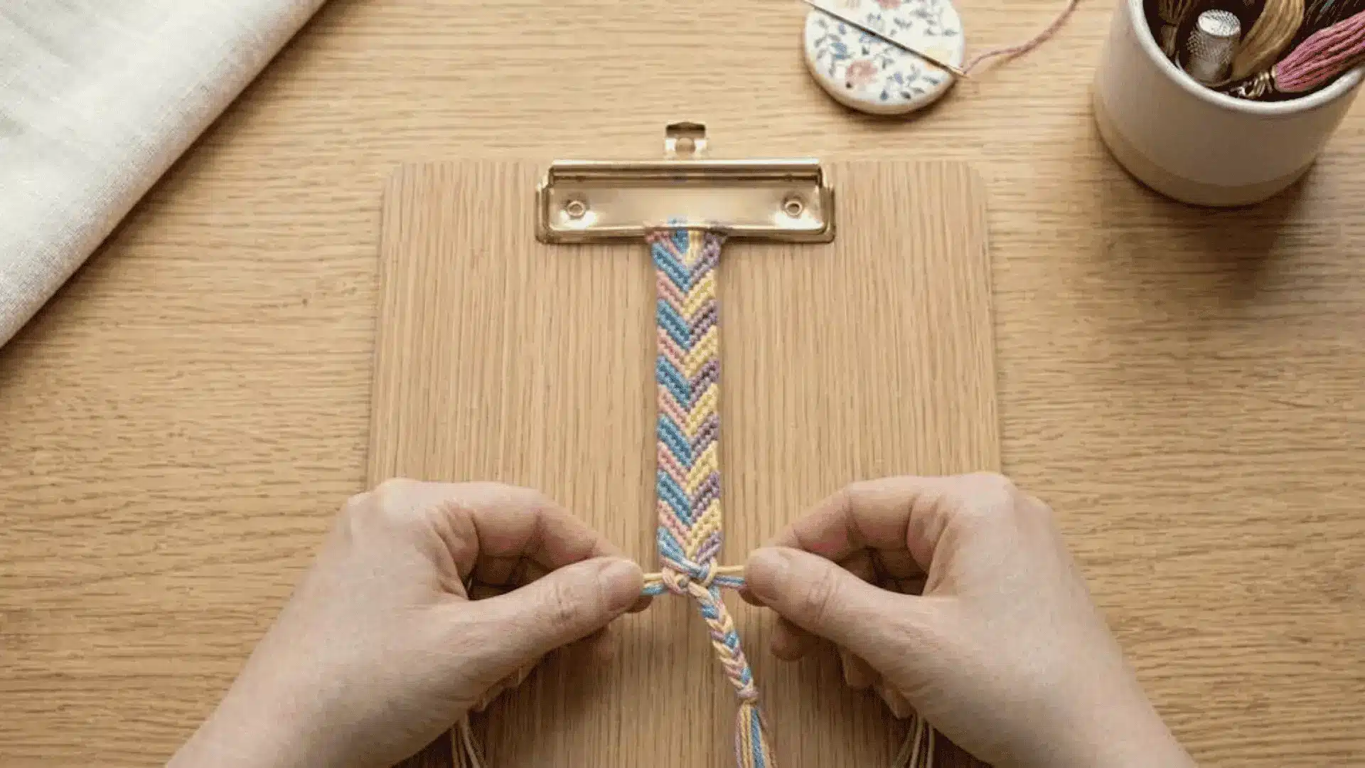 Hands tying the final knot on a completed embroidery floss friendship bracelet with a chevron pattern.