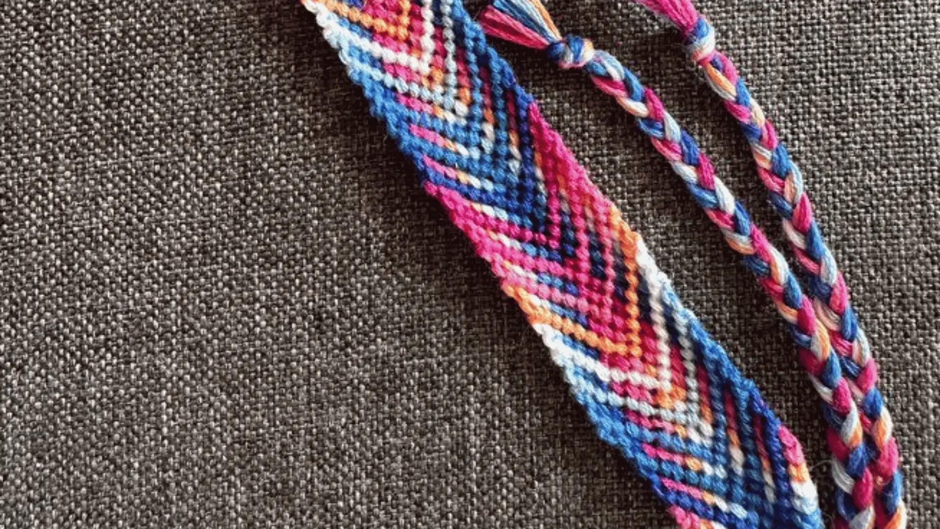 A vibrant, multicolored woven friendship bracelet with a fishbone pattern resting on textured grey fabric.