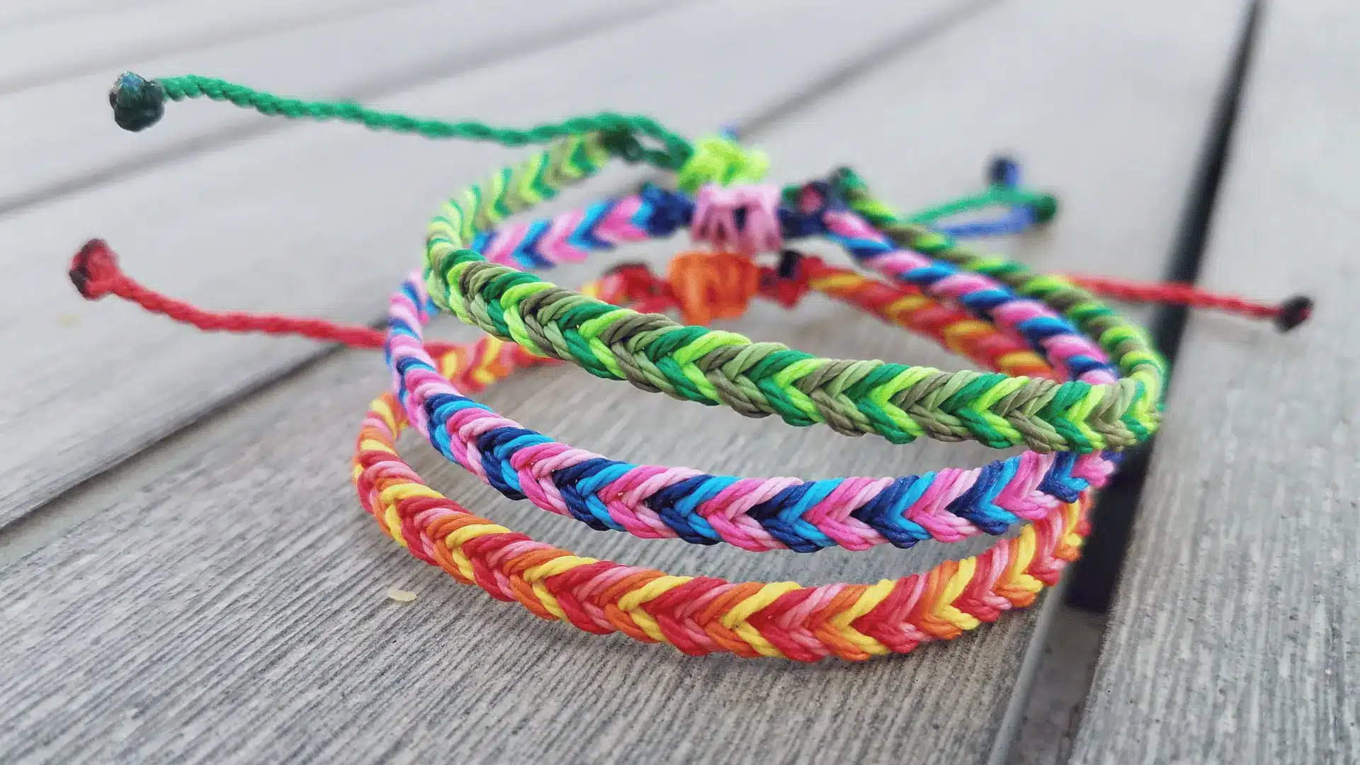 A stack of colorful, braided fishtail bracelets in green, blue, pink, and orange tones resting on a wooden surface.