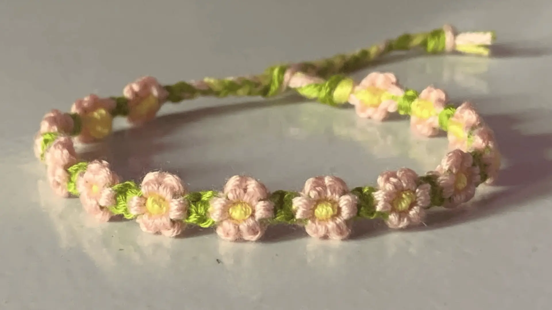 A dainty woven friendship bracelet featuring a pink, yellow, and green daisy floral stitch pattern.