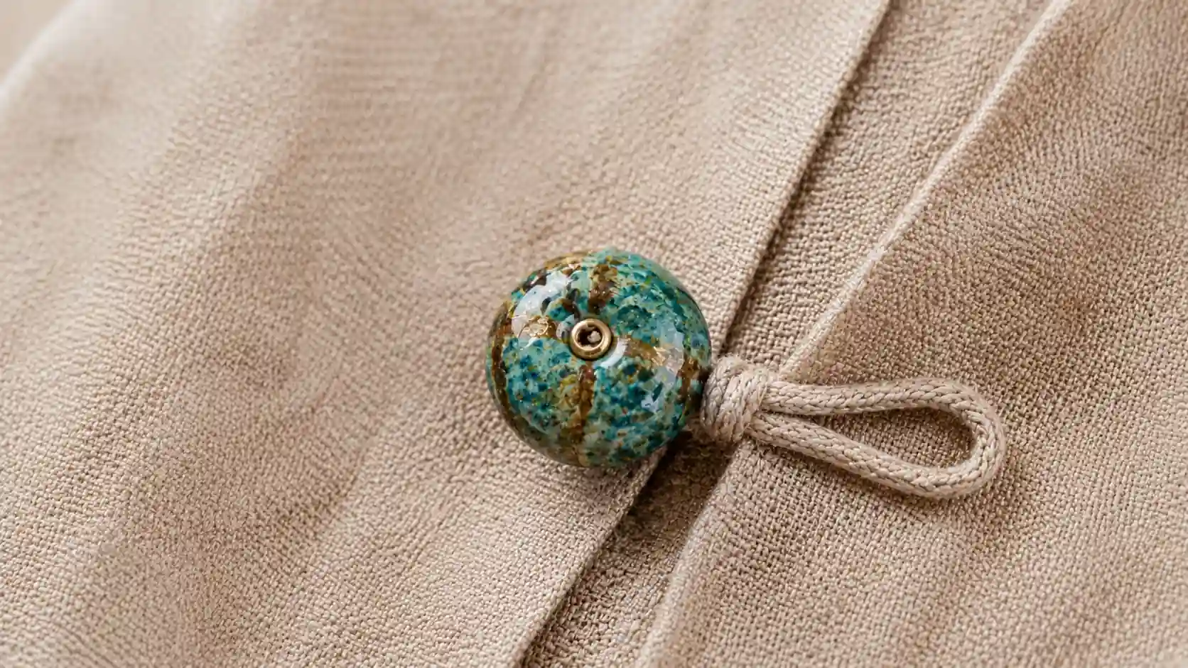 Turquoise bead button with cord loop closure on textured beige fabric in a handmade style.