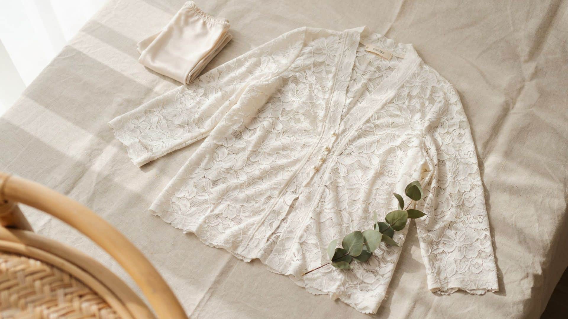 White lace cardigan with floral embroidery, scalloped hem, and button-front design styled on a soft neutral fabric background with eucalyptus accent.