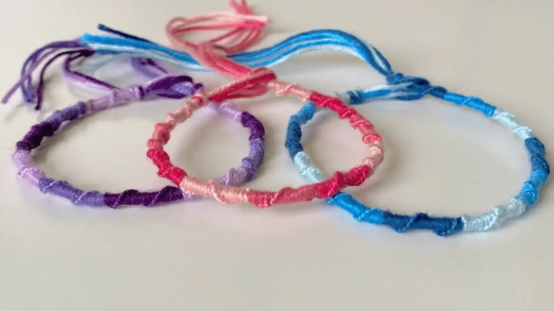 Three ladder-style friendship bracelets in shades of purple, pink, and blue laid out on a white surface.