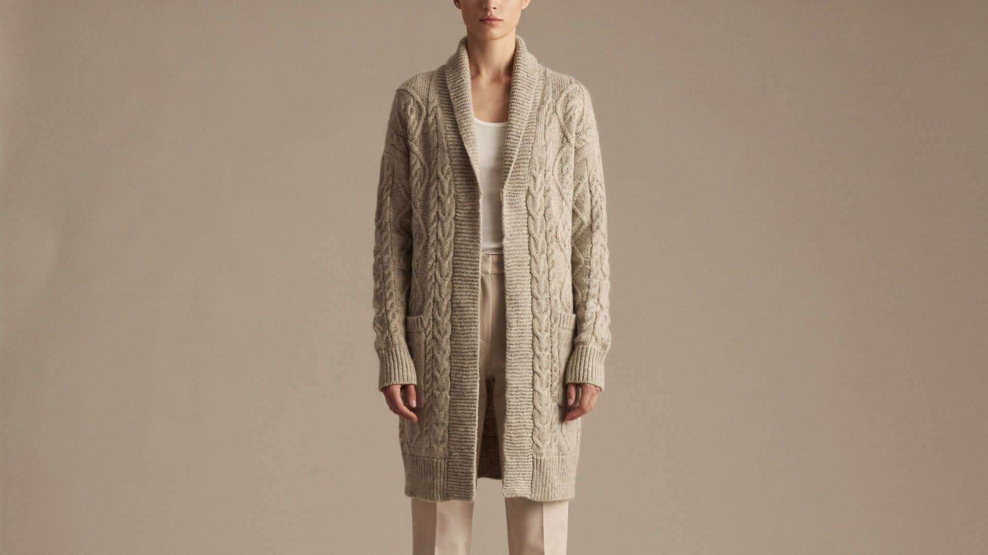 A longline cardigan in a neutral beige tone, featuring an open front, ribbed trim, and a relaxed oversized fit that falls below the knee, styled over a white fitted top and straight-leg jeans.