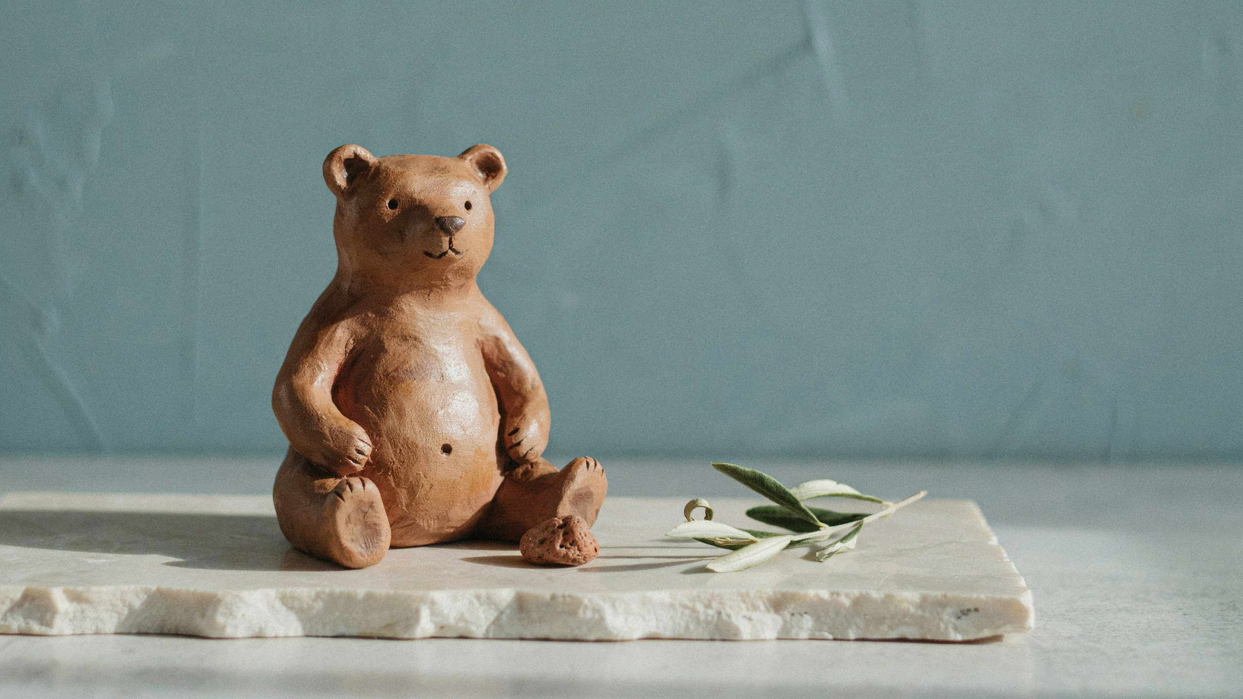 Brown and caramel clay bear figurine on stone surface with teal background, realistic handmade texture and soft natural lighting.