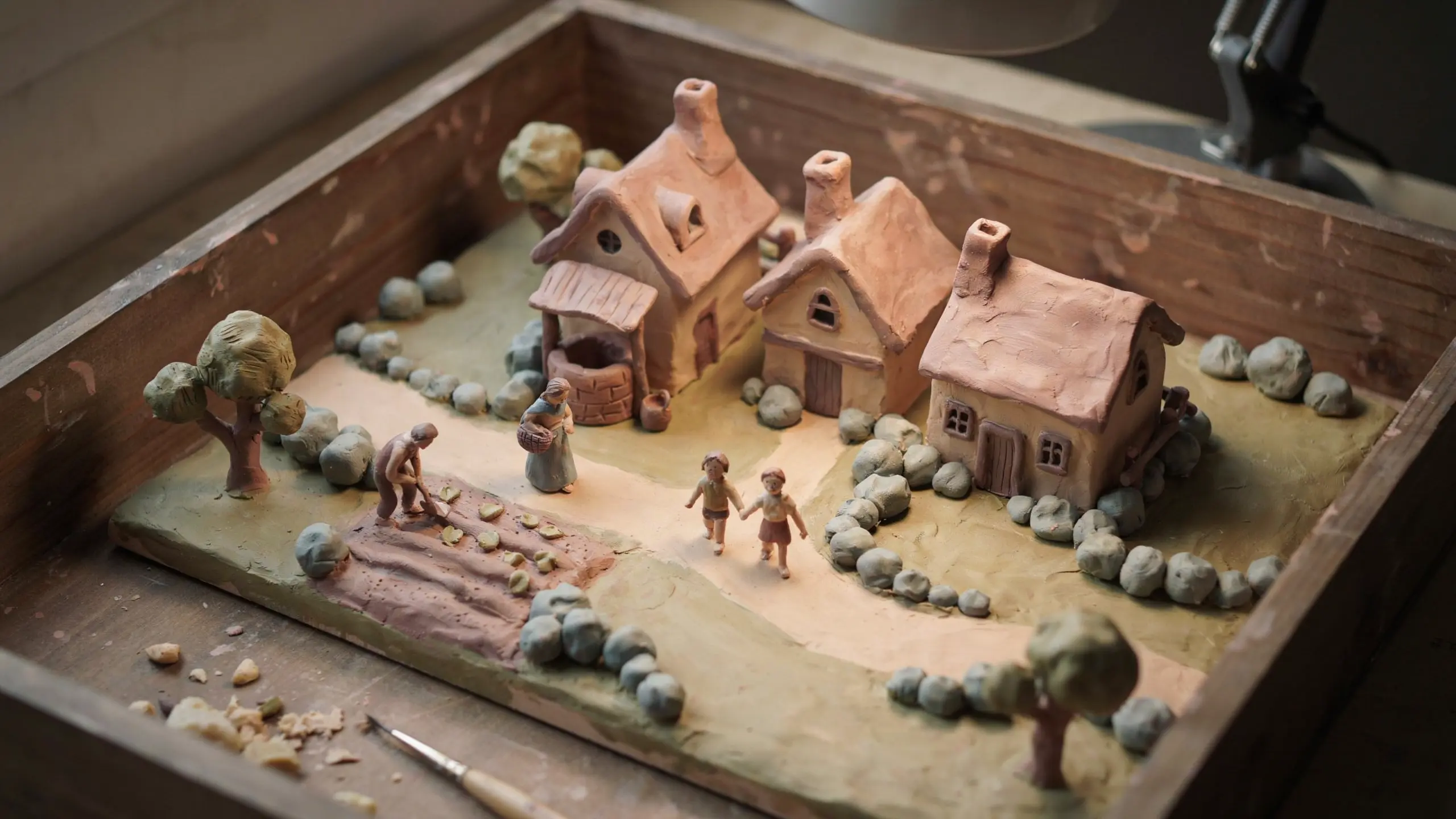 Handmade miniature clay diorama in a shallow box showing a tiny village scene.
