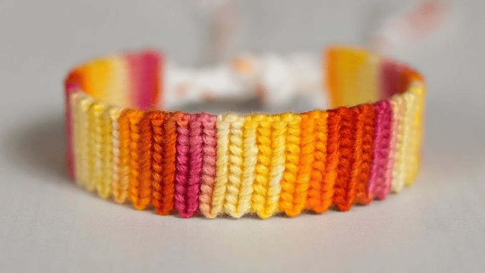 A woven ombre string bracelet with vertical stripes of yellow, orange, and pink thread on a plain background.