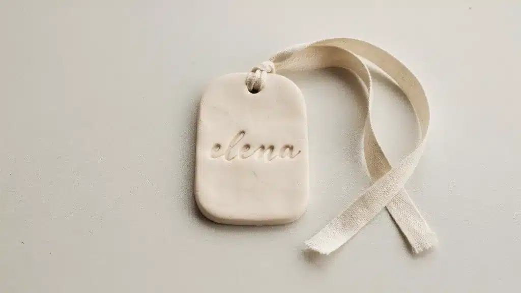A handmade polymer clay gift tag lying flat on a neutral table surface with engraved text, small hole detail, and ribbon placed beside it under soft lighting.