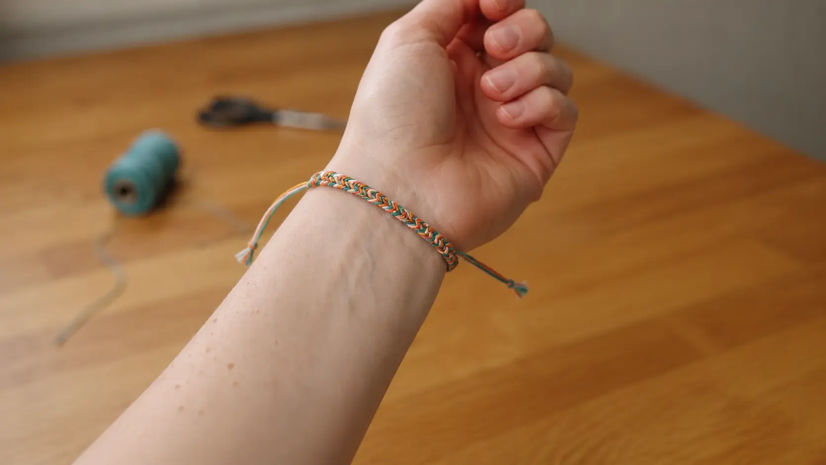 Hands placing a colorful braided bracelet around the wrist on a wooden crafting surface.