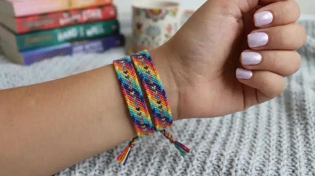 A wrist wearing two colorful woven rainbow chevron friendship bracelets, with books and a mug blurred in the background.