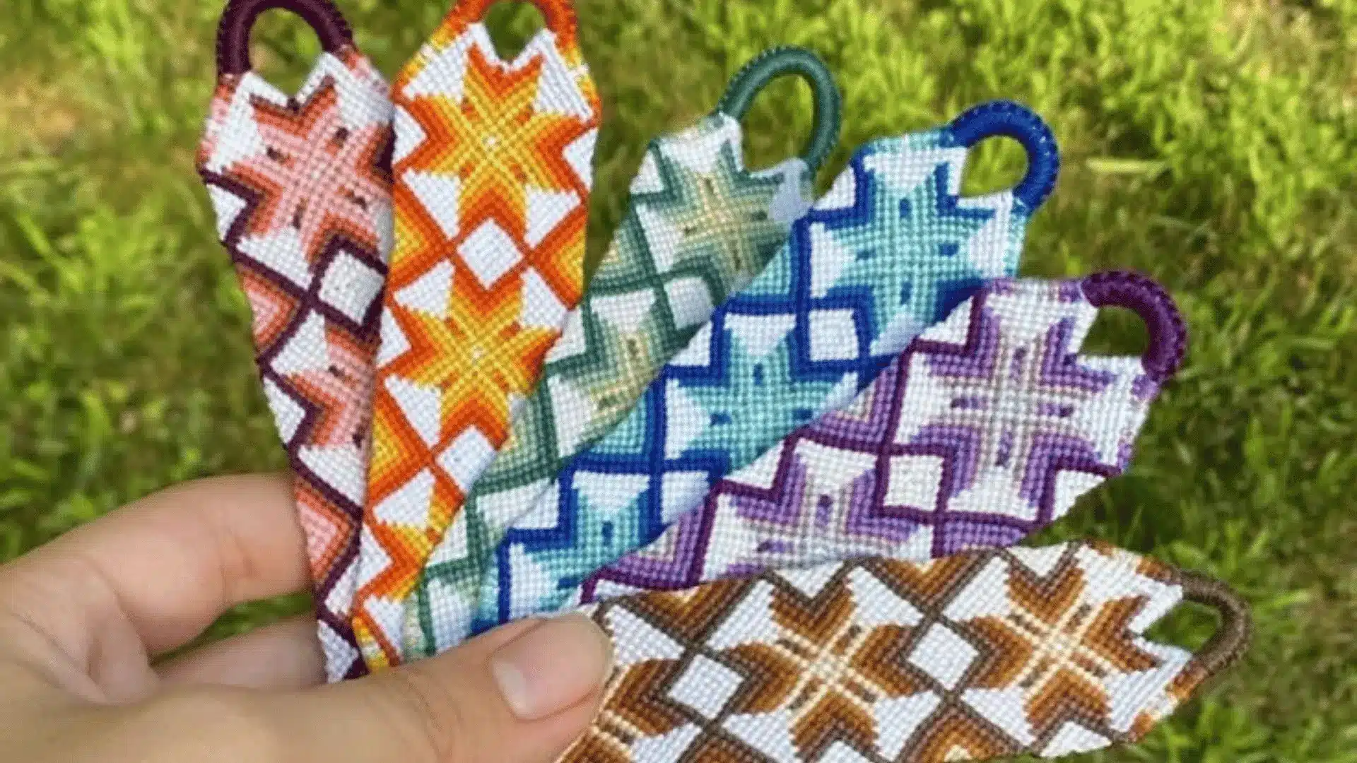A hand fanning out six woven friendship bracelets featuring a colorful starburst design against a grassy background.