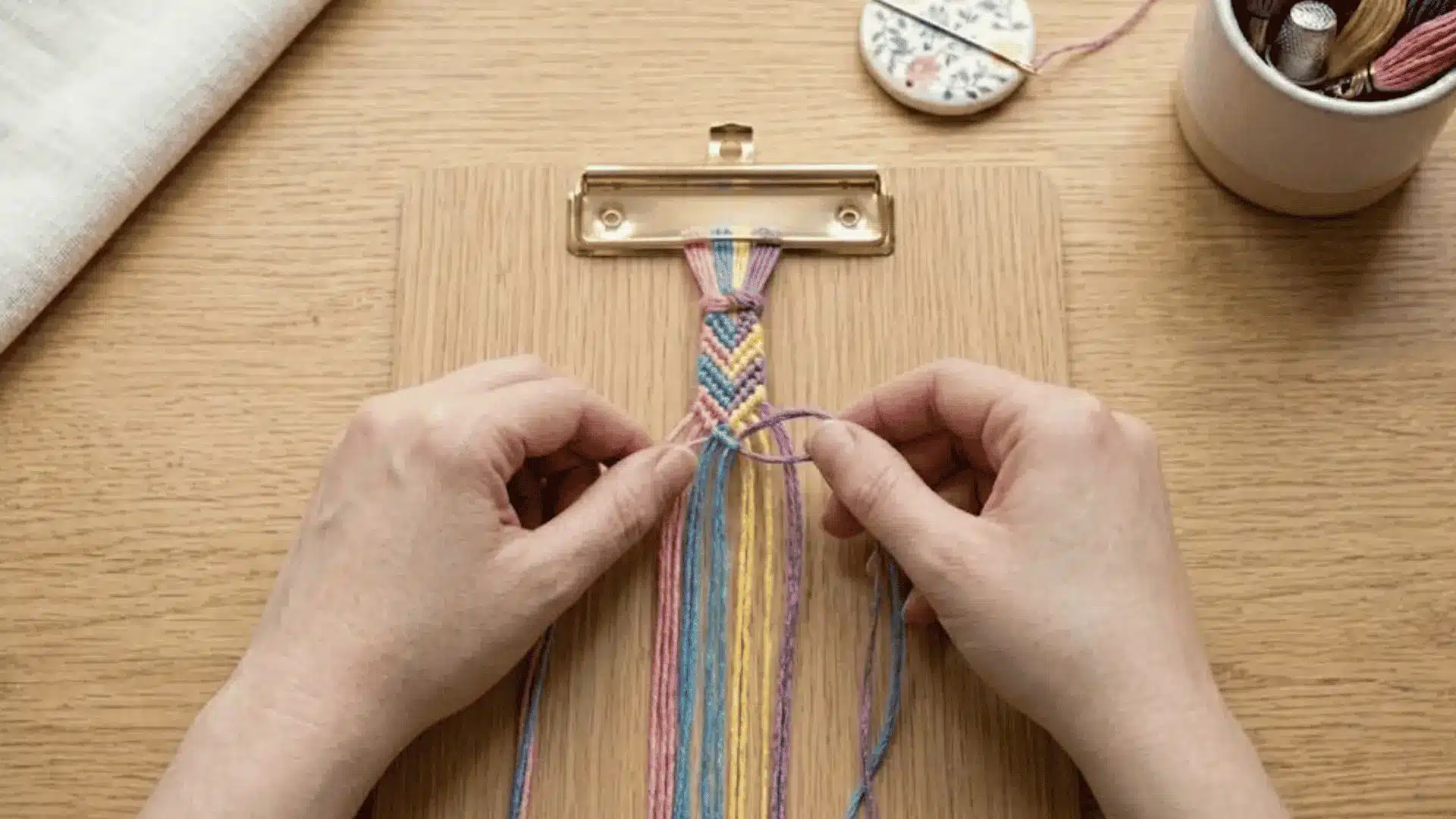 Hands tying the first knots of an embroidery floss friendship bracelet attached to a clipboard.