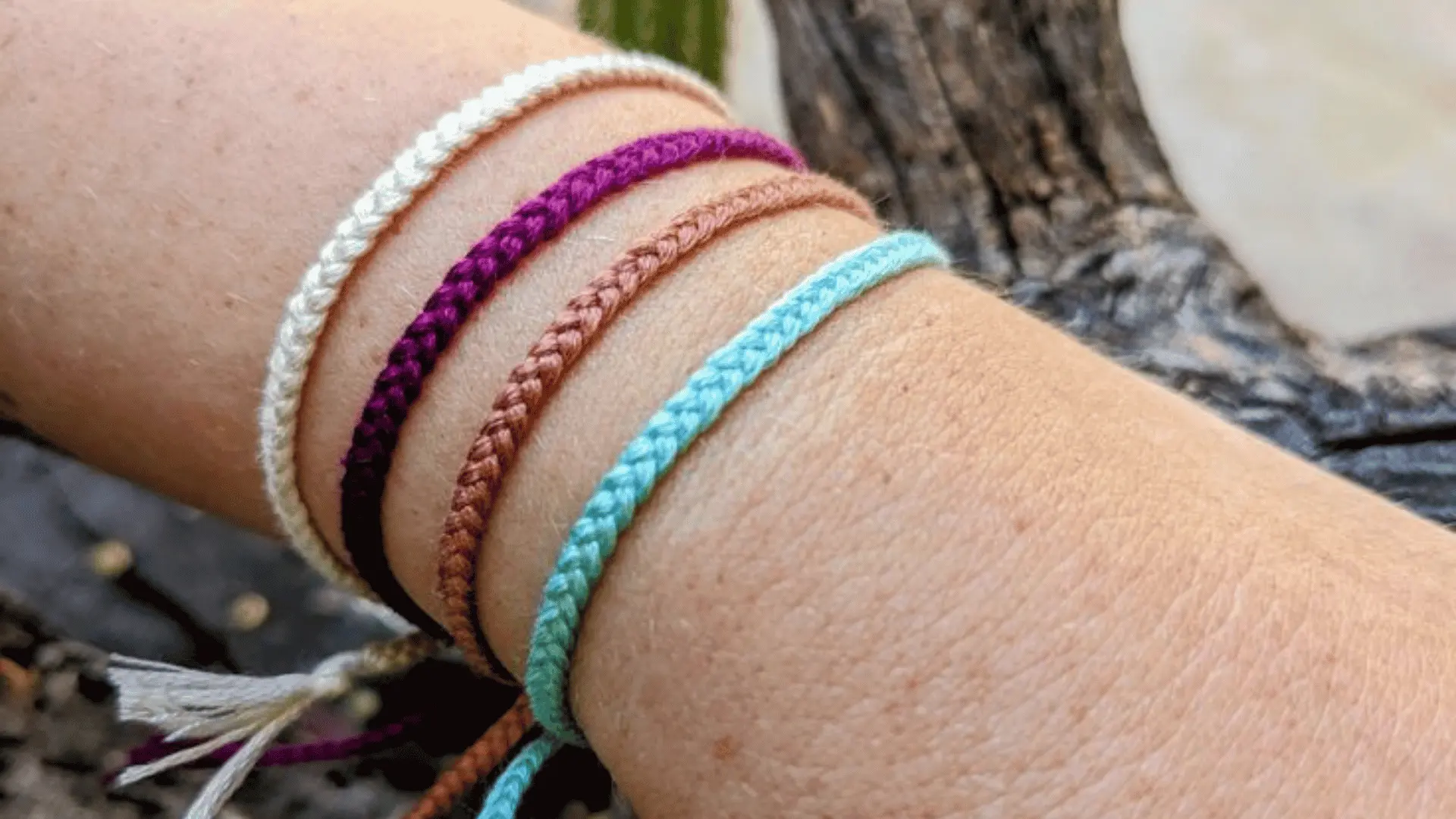 Four thin line minimal braided friendship bracelets in cream, purple, brown, and light blue stacked on a wrist.