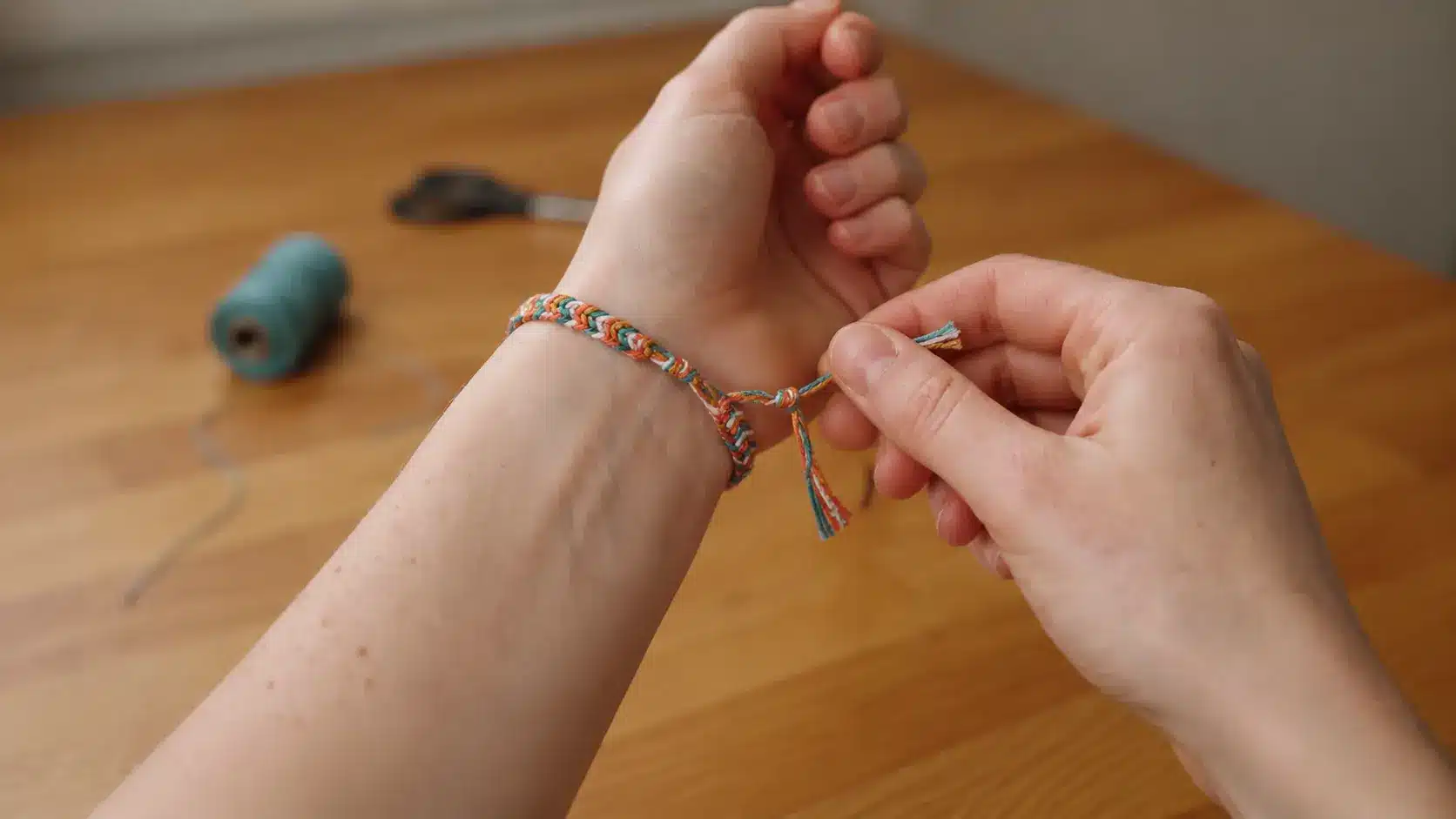 Hands tying the braided ends of a colorful friendship bracelet over a wooden surface.