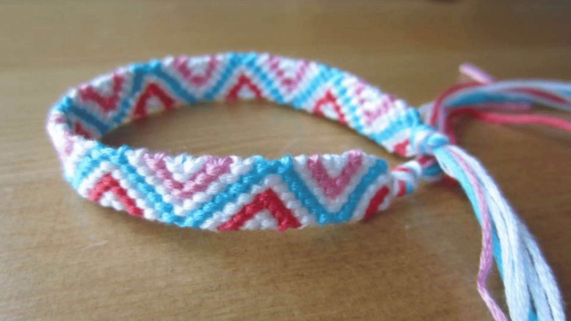 A woven friendship bracelet with a red, pink, blue, and white triangle chain pattern resting on a wooden surface.