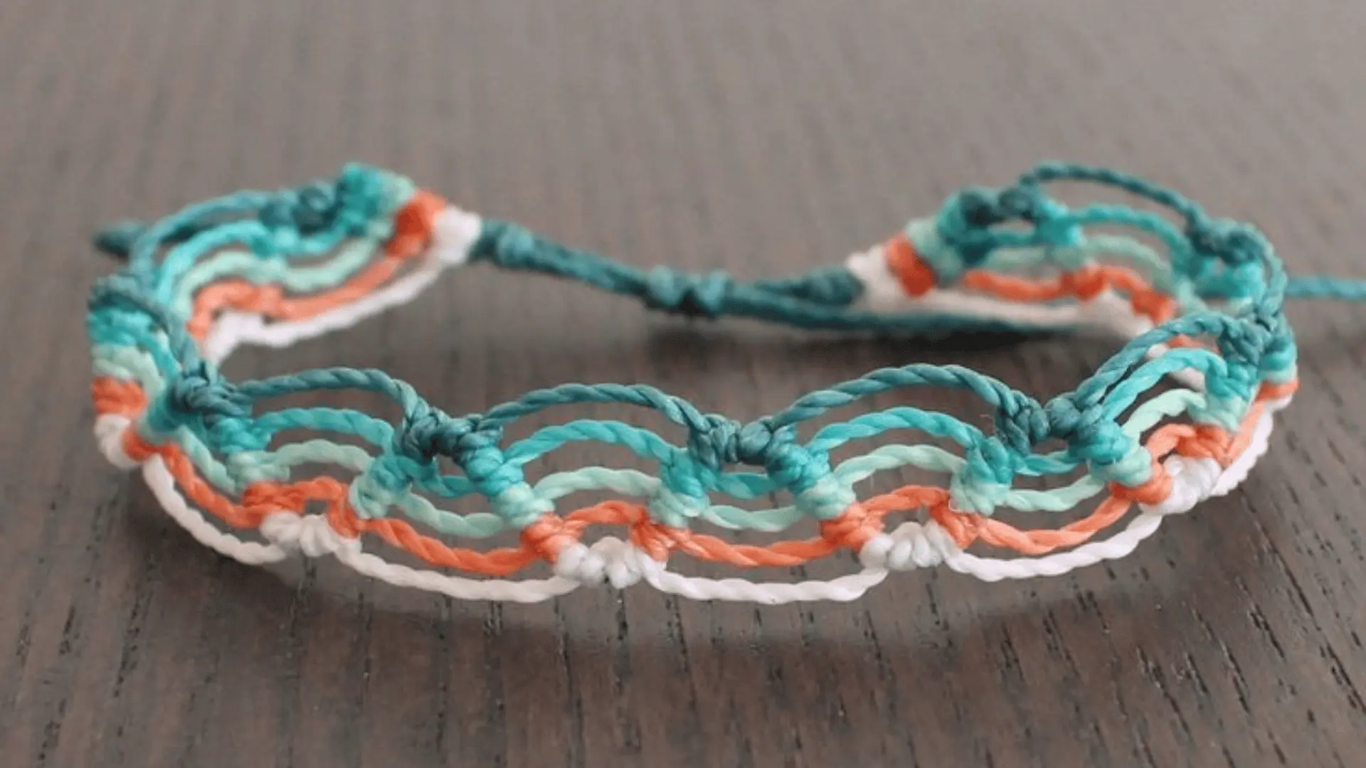 A teal, orange, and white wave beach macrame friendship bracelet resting on a dark wooden surface.
