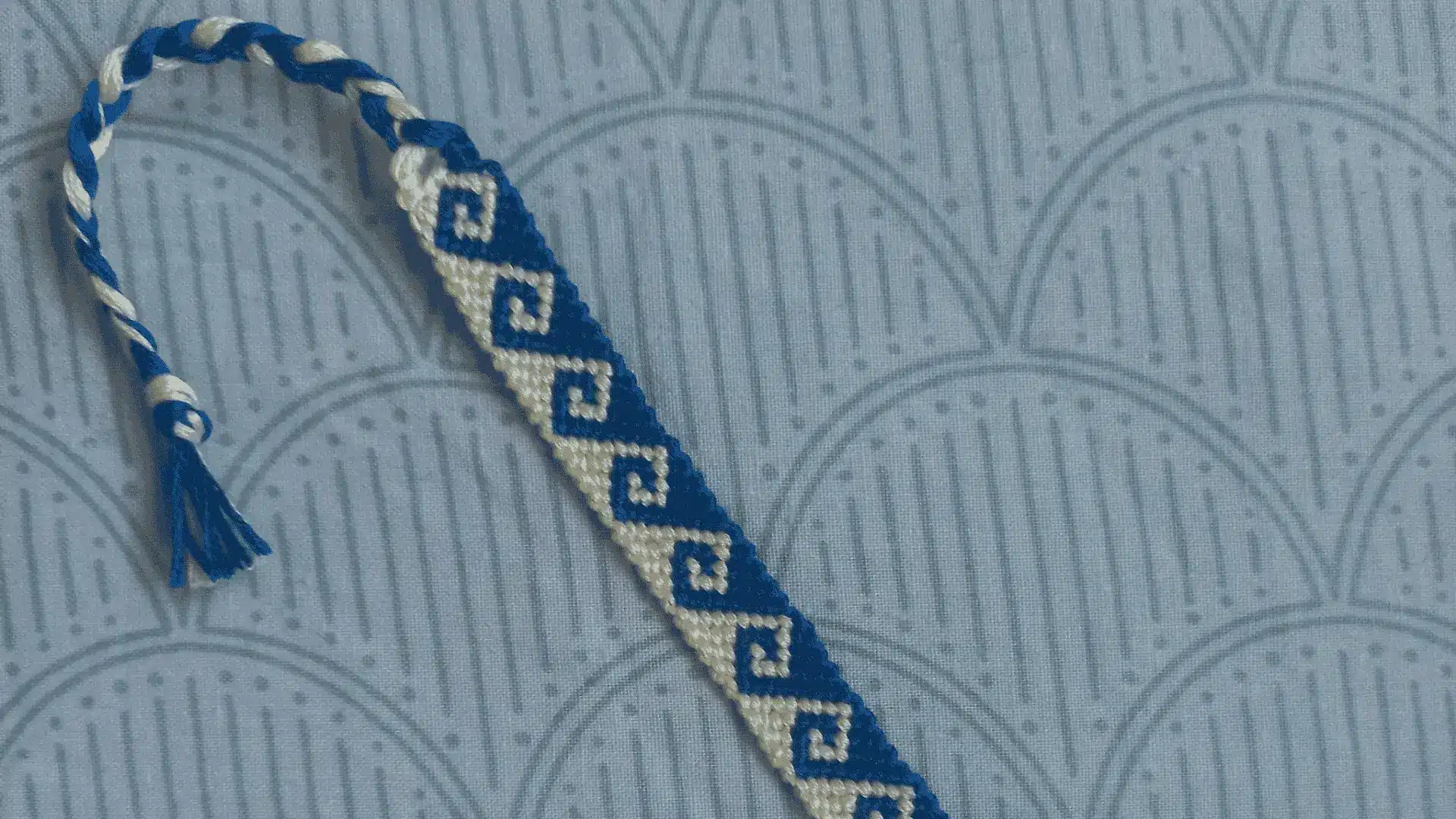 A blue and white woven friendship bracelet with a wave pattern resting on a patterned blue fabric.
