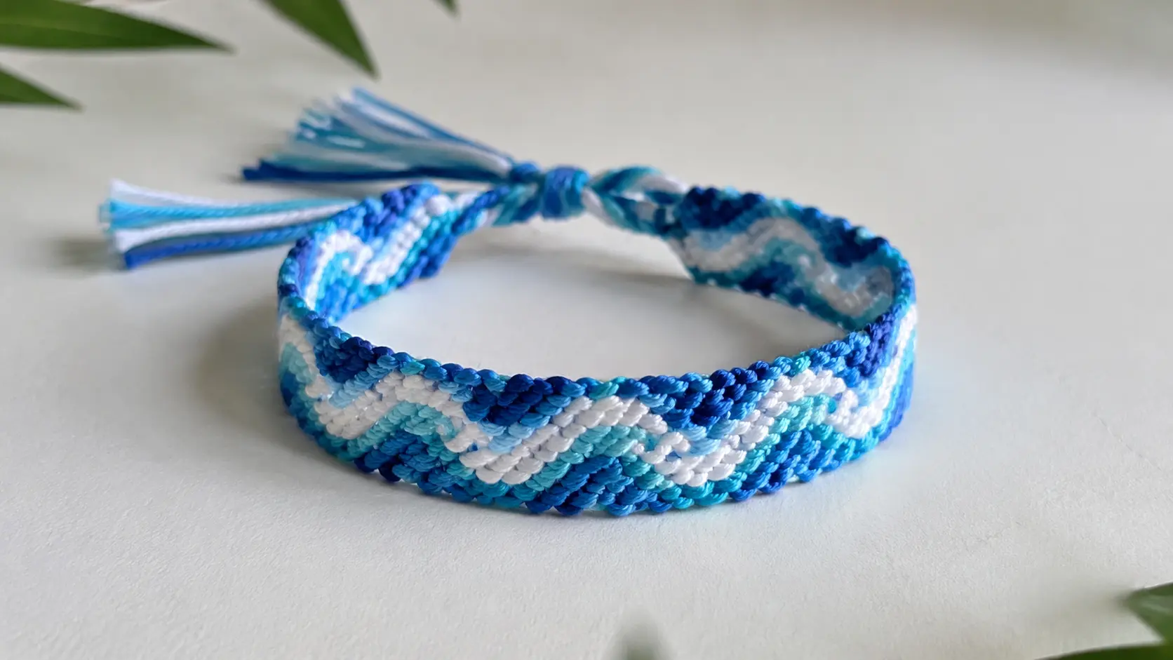 Blue and white wave friendship bracelet with braided tassels, shown in close-up on a neutral surface.