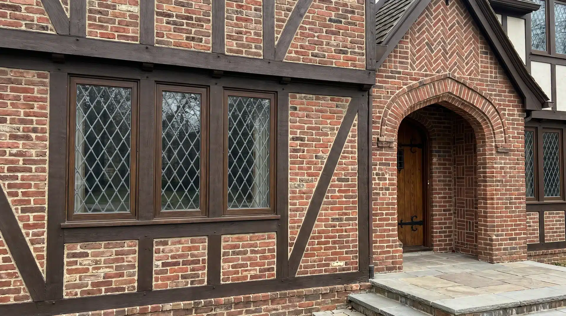 Red brick and dark timber-framed house exterior with diamond-paned windows, an arched brick doorway, and stone paving at the entrance.