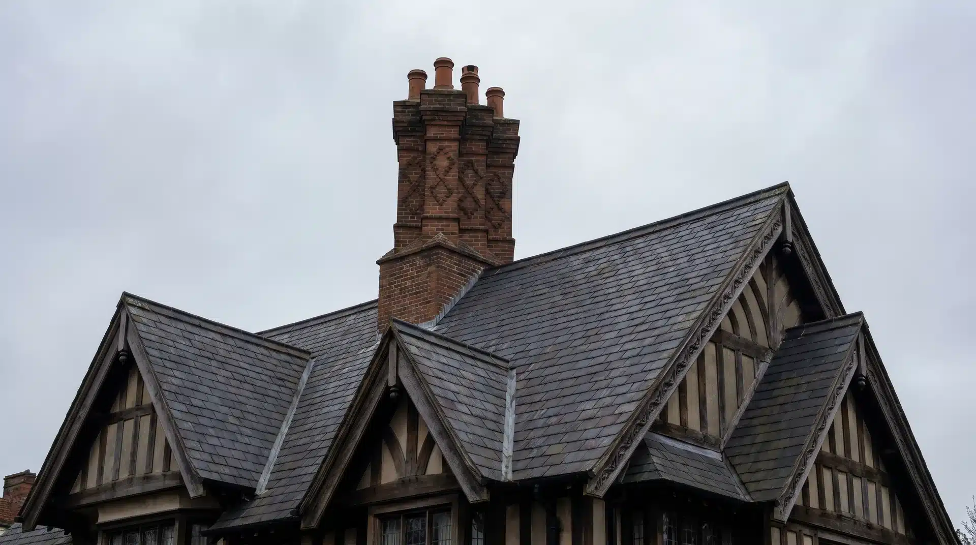 Dark slate roof with timber-framed gables and a brick chimney on a traditional house under an overcast sky.